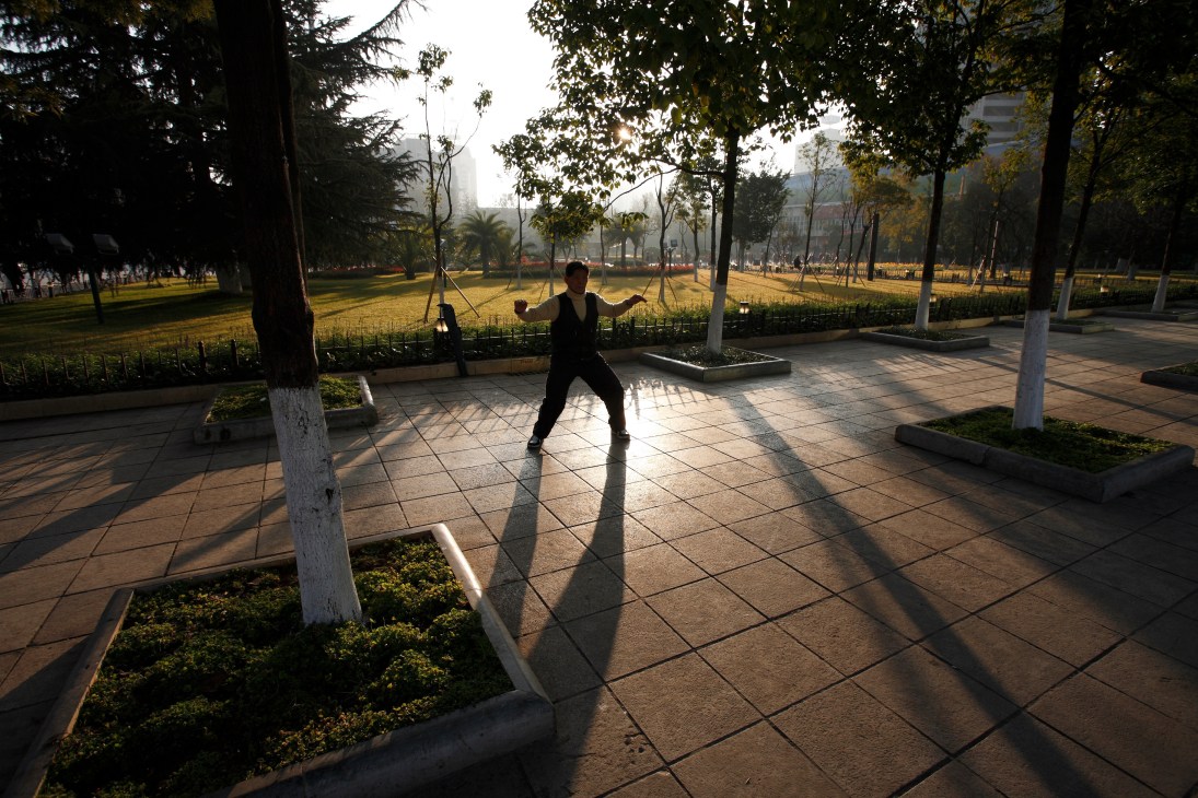 A person in dark clothing exercises in Dong Feng Square at sunrise, surrounded by trees and manicured landscaping.