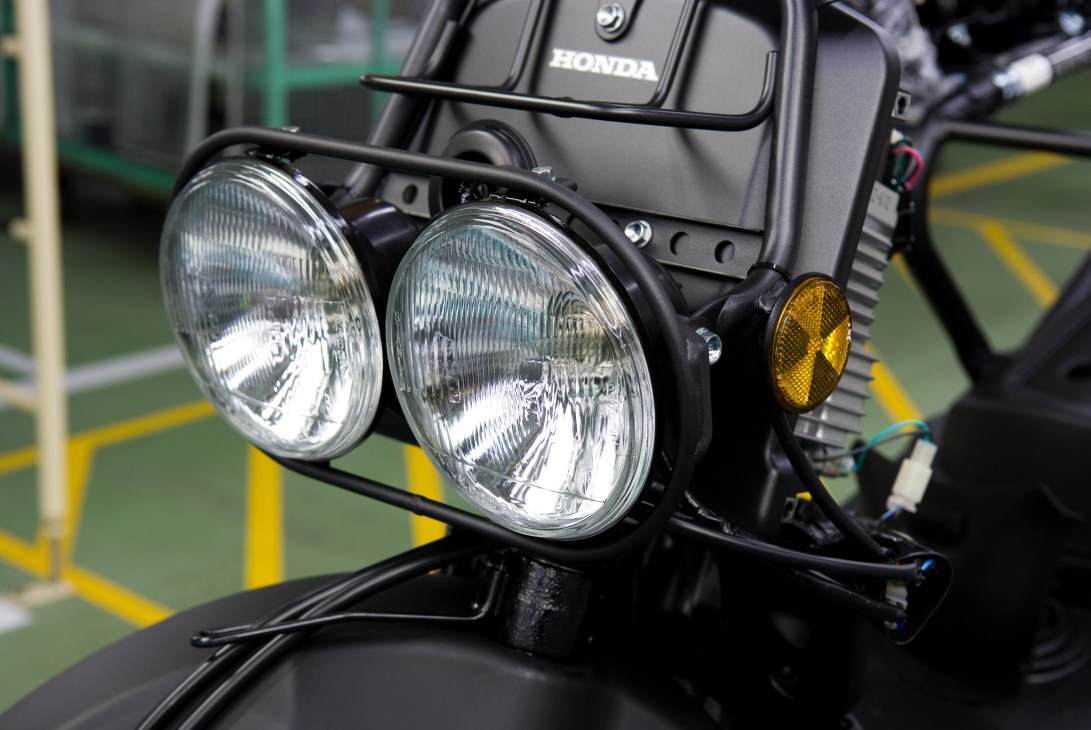 Close-up of a Honda Zoomer's distinctive dual round headlights with chrome bezels and yellow indicator light.