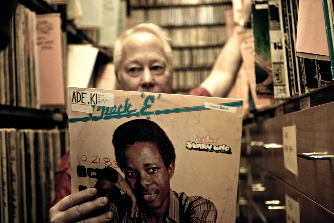 Tom Schnabel holds a Chuck E record in KCRW's music library surrounded by shelves of vinyl and CDs.
