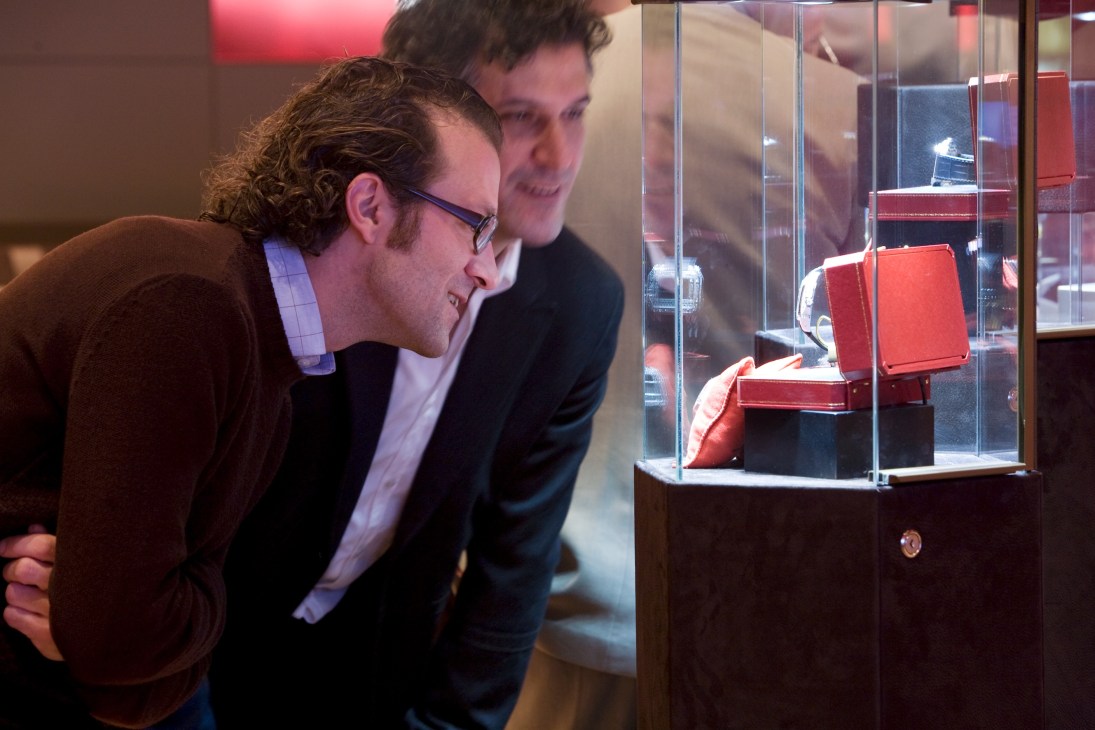 Two men in business attire examine red luxury watch boxes displayed in a glass case at Cartier.