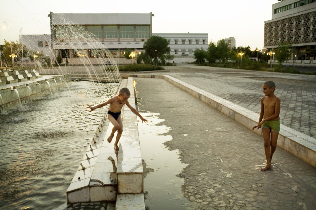 Two shirtless boys play in a shallow Soviet-era fountain with water sprays in a public plaza surrounded by modern government buildings.