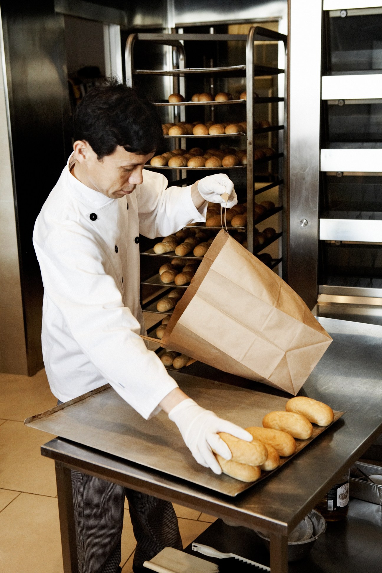 Baker in white chef's coat packaging fresh bread rolls into a paper bag at Andersen bakery.