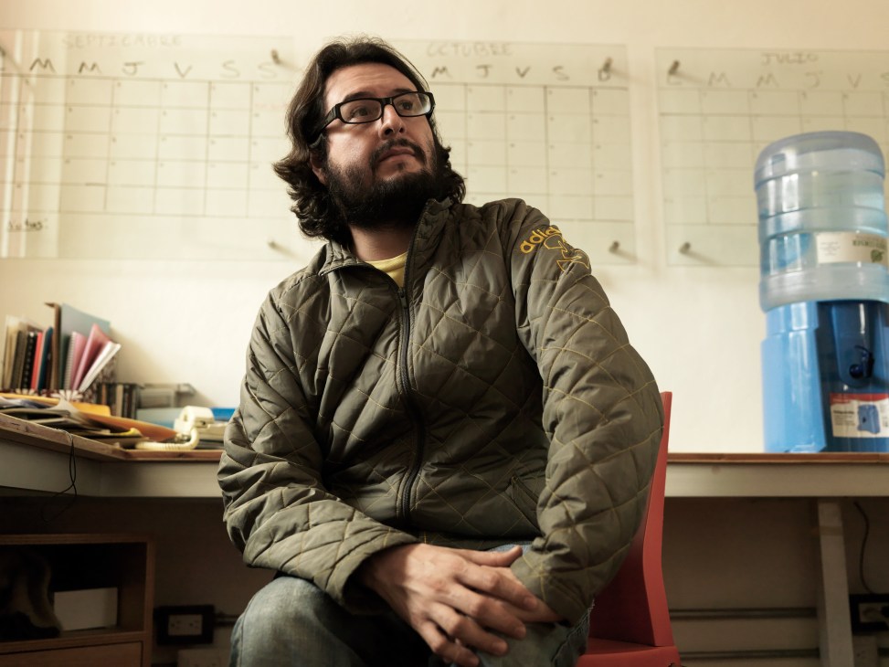 Producer Pablo Cruz wearing glasses and a quilted jacket sits in an office with scheduling charts and office supplies visible behind him.