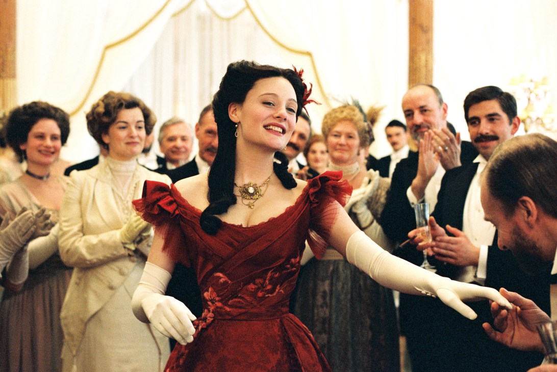 Woman in red dress at formal ball surrounded by guests in elegant period attire.