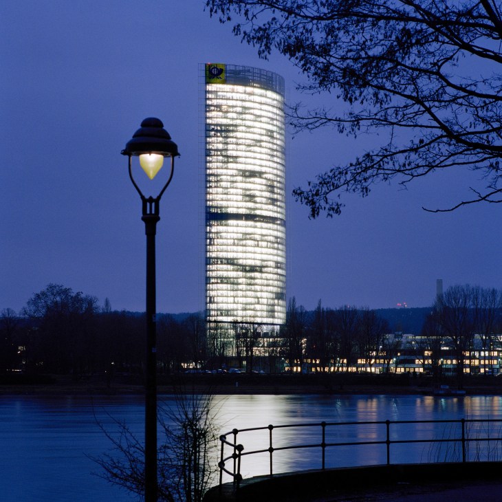Illuminated cylindrical office tower reflected in water at dusk with street lamp in foreground.
