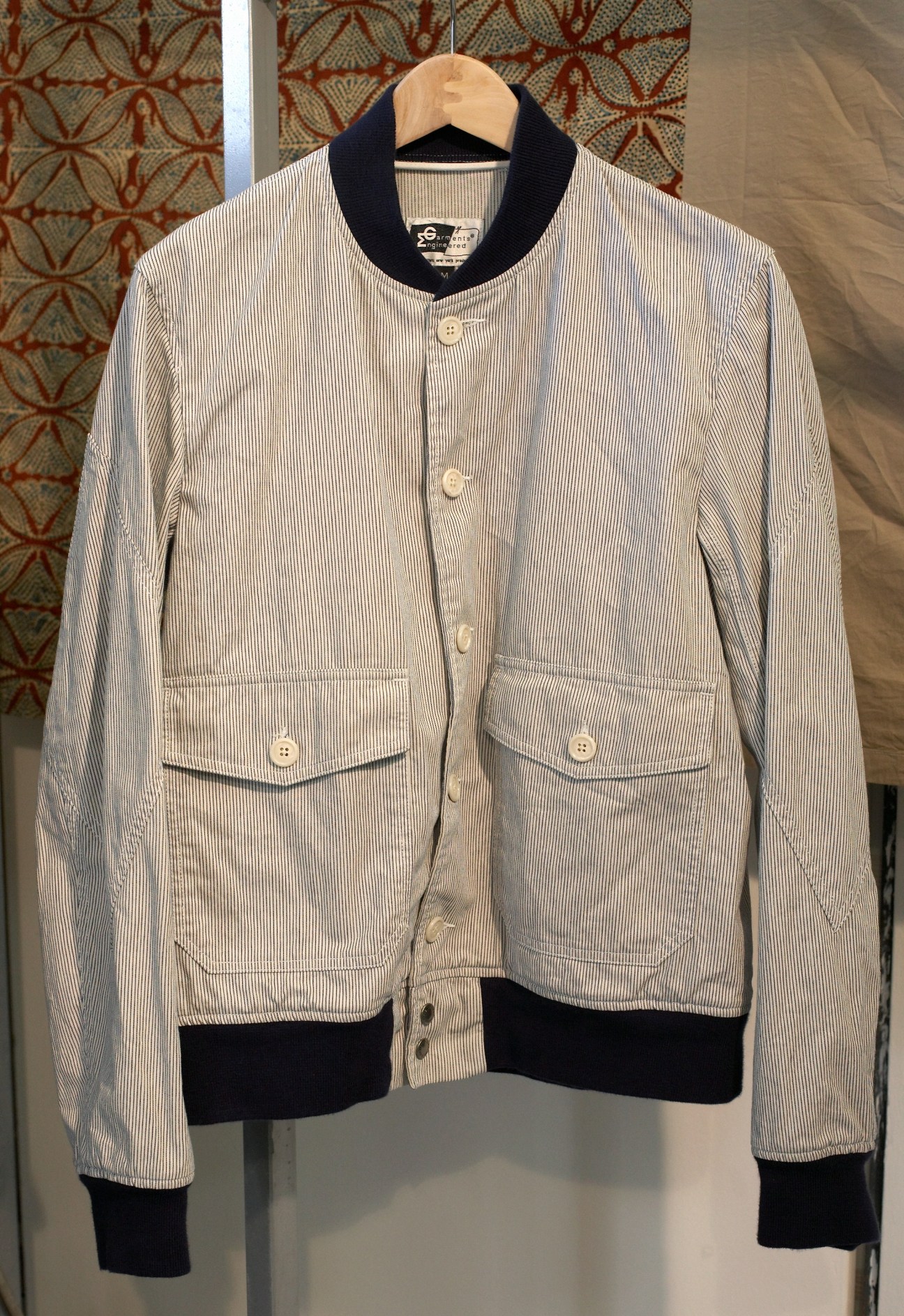 Striped light-coloured jacket with black collar and cuffs on wooden hanger against patterned backdrop.
