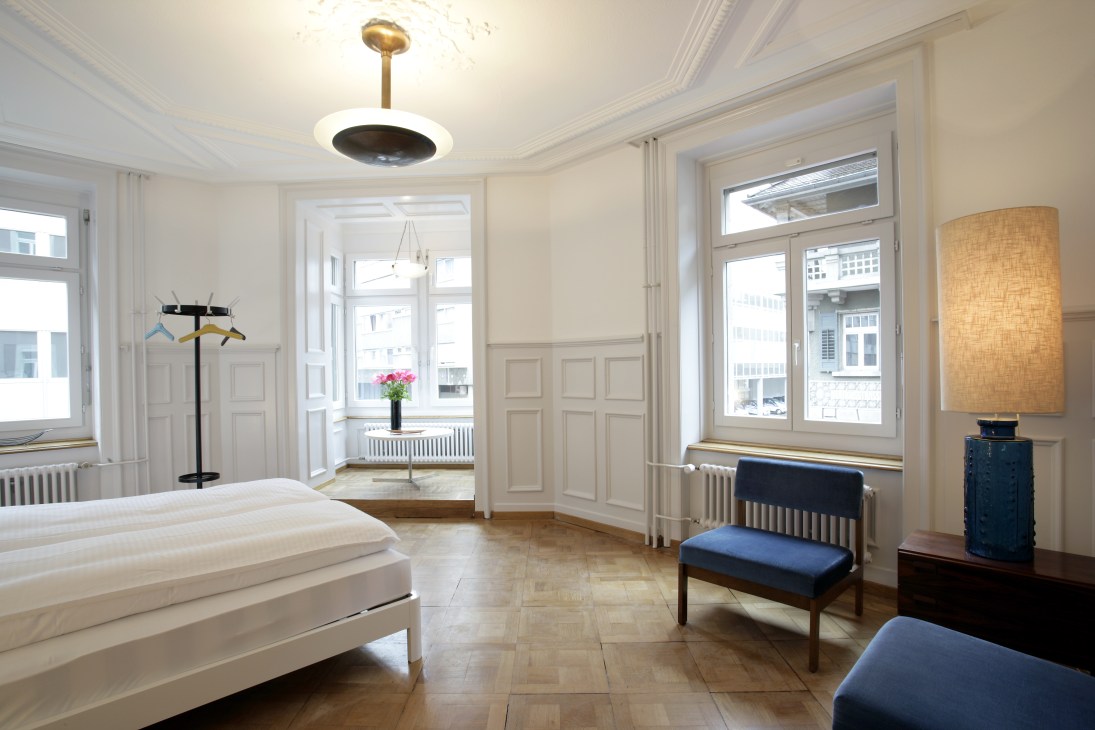 Bright hotel bedroom with white bed, blue bench seating, and large windows overlooking a city street.
