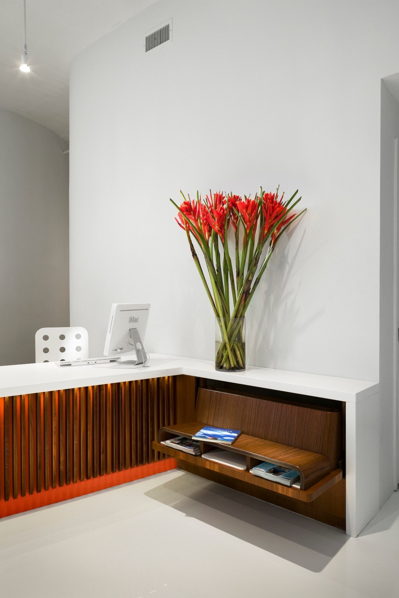 Modern office reception desk with white lacquer top, walnut storage, orange accent panel and red flowers in a vase.