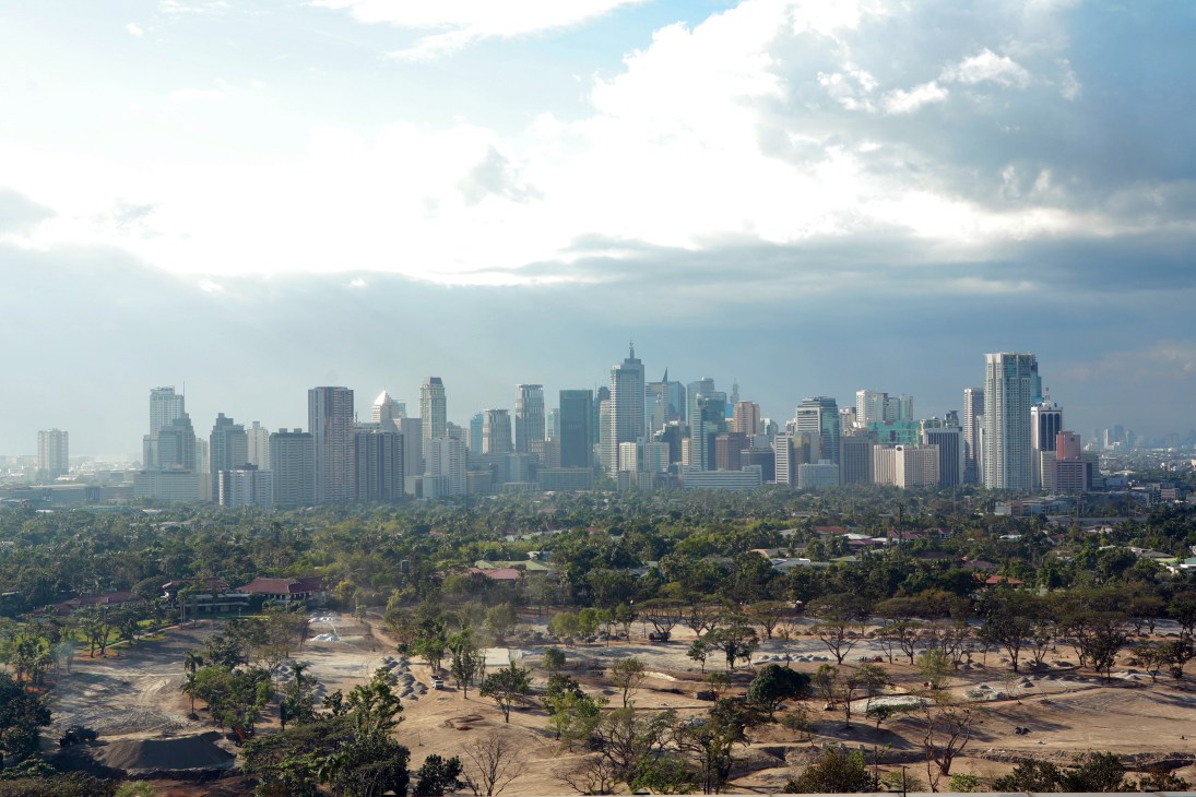 Makati skyline with tall office buildings rising above low-rise residential areas and green vegetation under a cloudy sky.