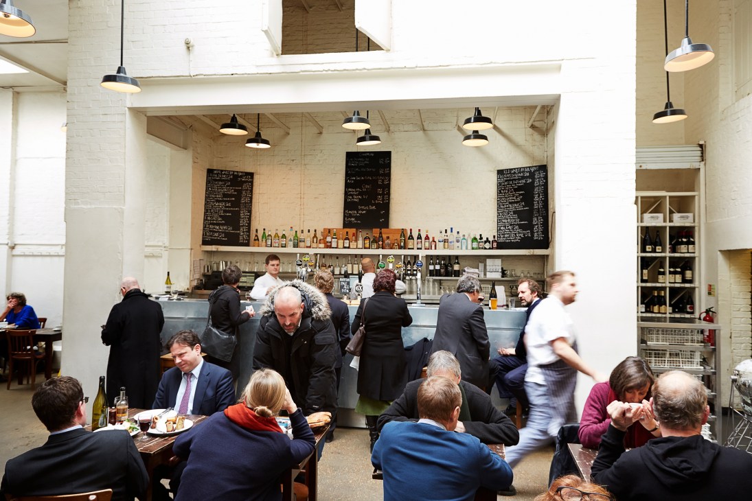Interior of St John restaurant in Smithfield showing diners at tables, bar counter with bottles and blackboard menus, and pendant lighting.