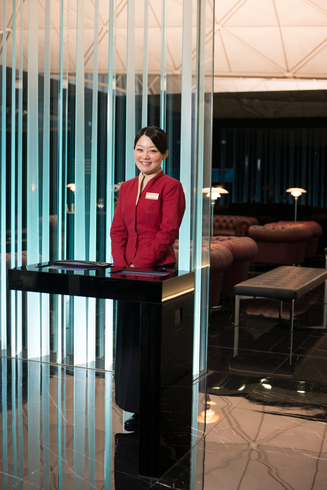Cathay Pacific staff member in red uniform at reception desk in modern first-class lounge with glass partitions.