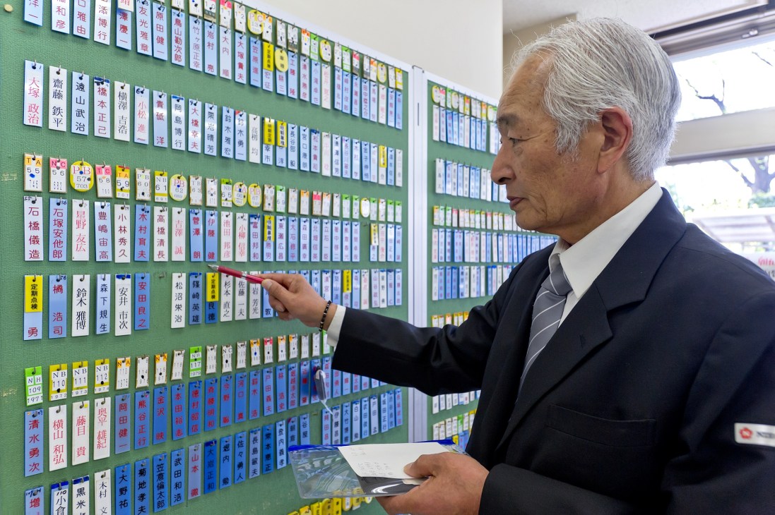 Man in dark suit pointing at wall of driver name plates displayed on green board