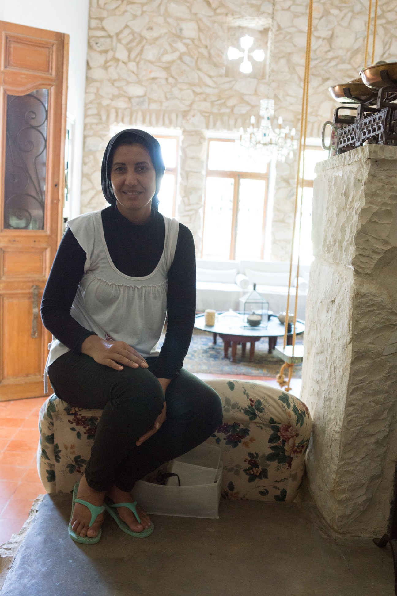 Woman wearing a black hijab and white apron smiling at camera in a stone-walled interior at Beit al Batroun hotel.