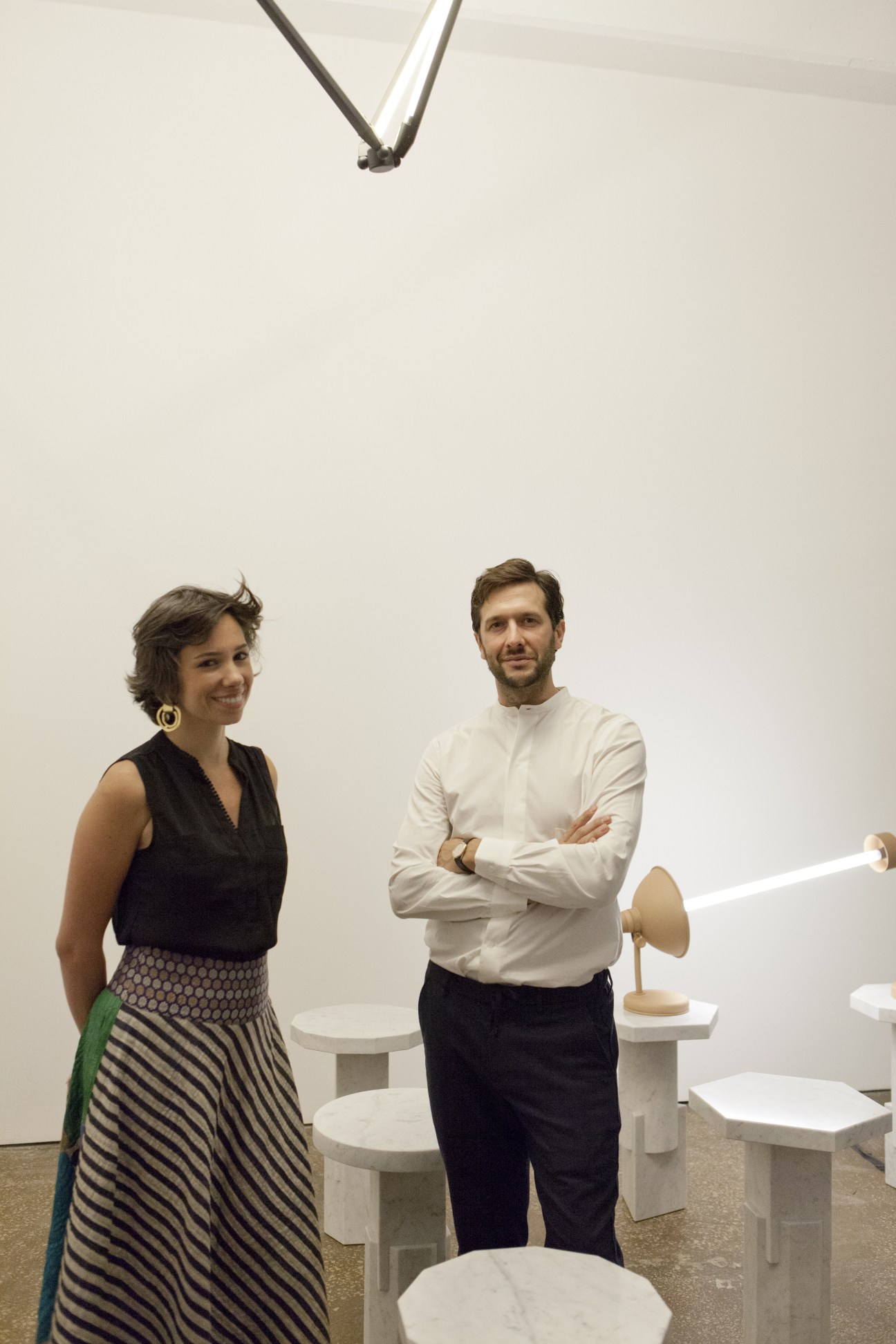 Pascale Wakim and Nicolas Bellavance-Lecompte stand in a gallery space with white furniture and pendant lights.