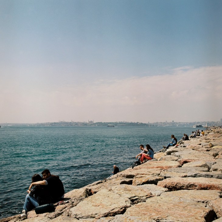 People sitting on rocky waterfront at Moda with Istanbul skyline across the water in the distance.