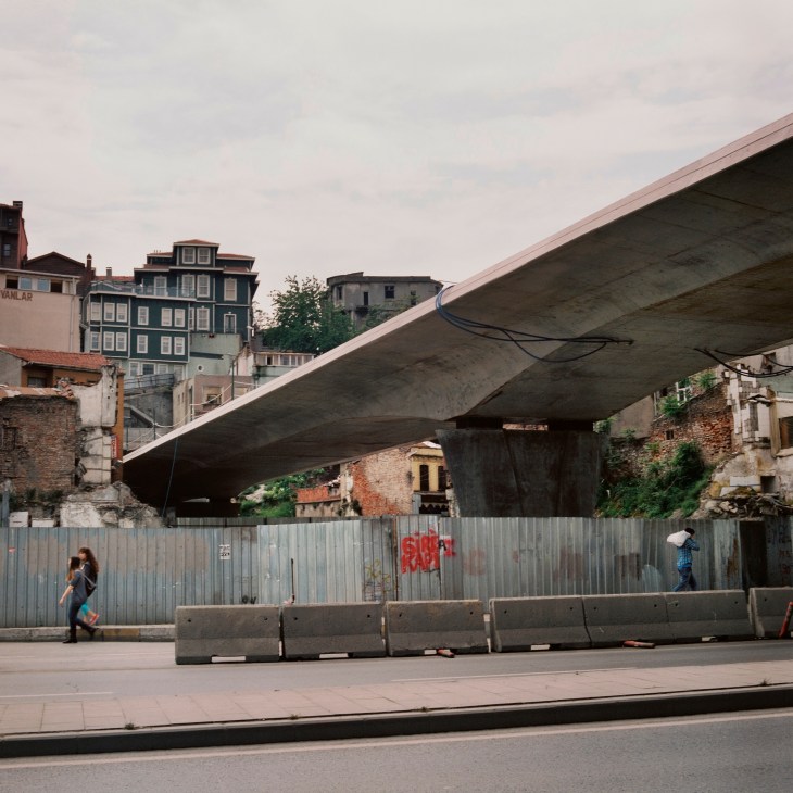 A concrete metro bridge spans over old Istanbul buildings and a street barrier with graffiti.