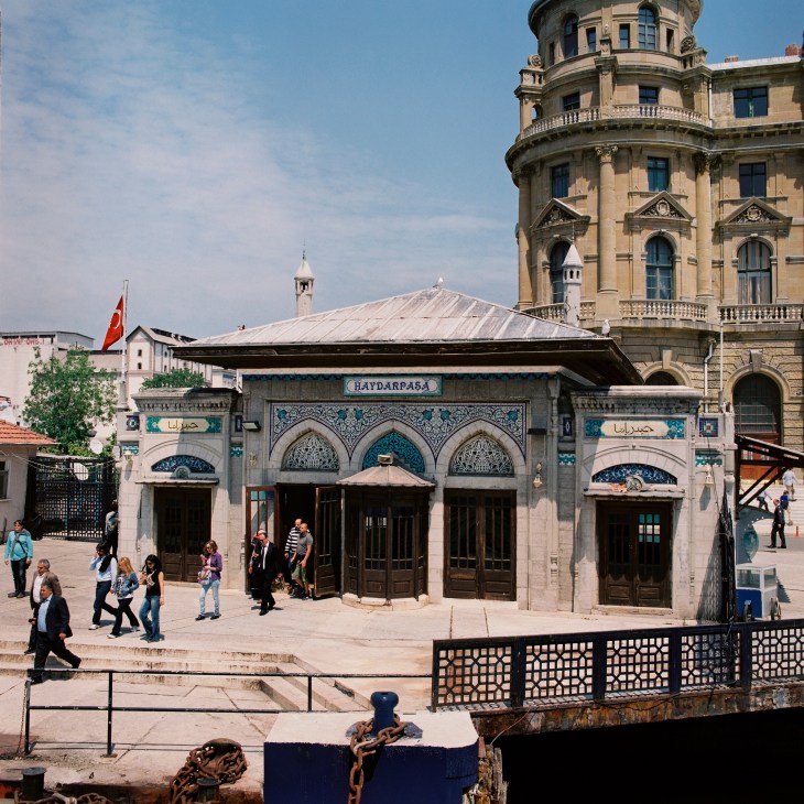 Haydarpasa station's ornate facade with blue tilework and arched doorways, with a historic building and Turkish flag visible behind it.