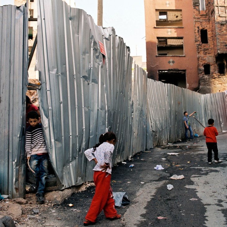 Children playing among corrugated metal barriers in a demolition site surrounded by damaged buildings in Istanbul.