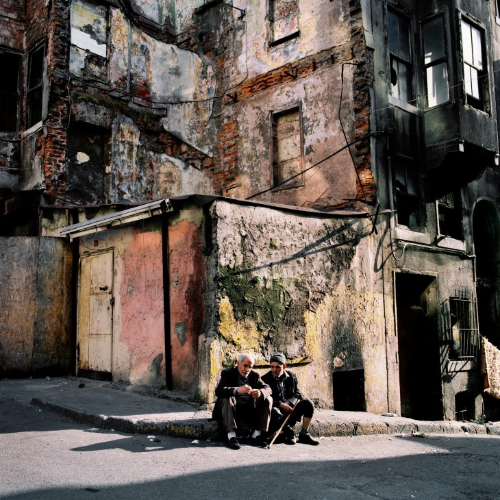 Two elderly men sit on a curb in front of a derelict building with crumbling walls and boarded windows in Tarlabashi, Istanbul.