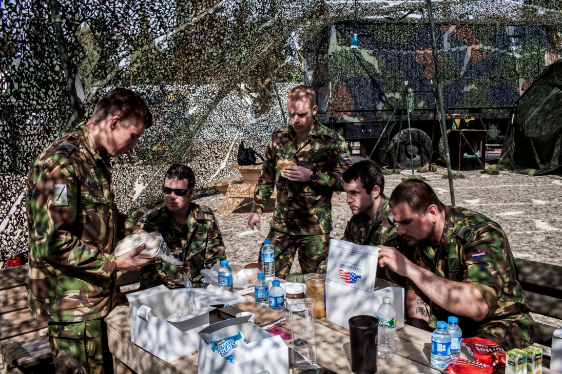 Dutch military personnel in camouflage uniforms eating lunch under camouflage netting at a Patriot missile base camp.