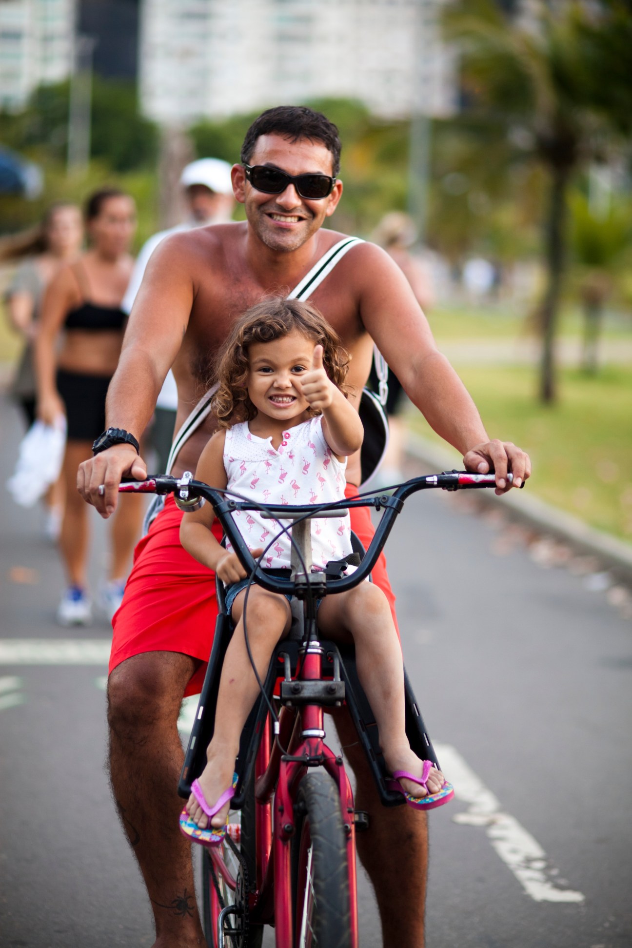 Man and young girl riding a red bicycle together on a Rio de Janeiro street.