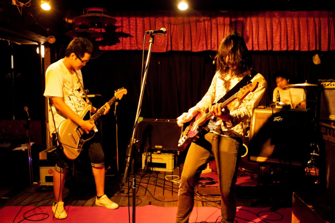 Musicians perform on stage with guitars under red stage lighting and curtains.