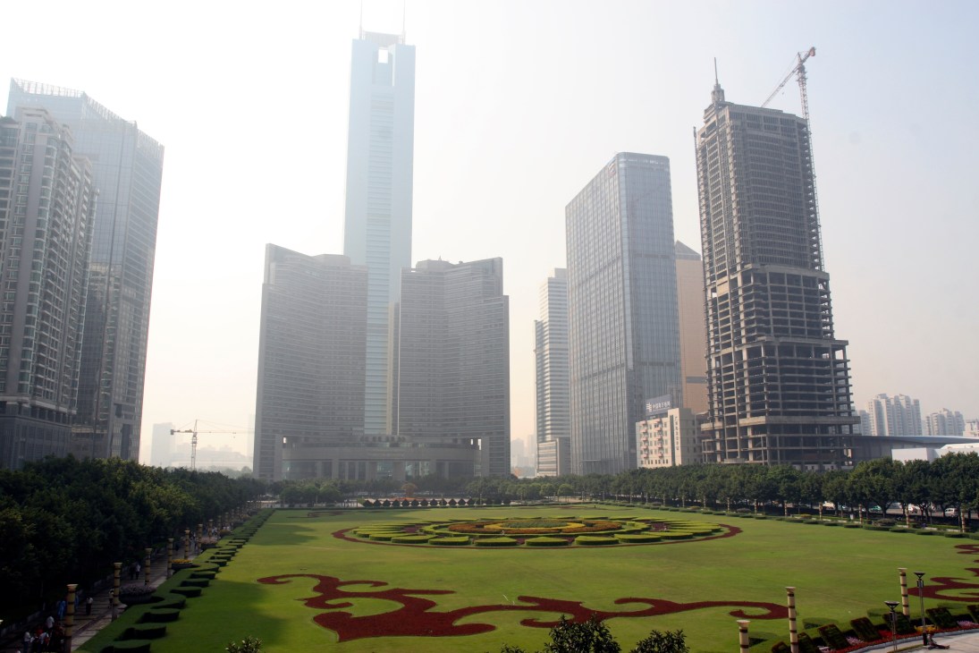 Modern skyscrapers surround a manicured green square with red landscaping in Guangzhou's financial district.