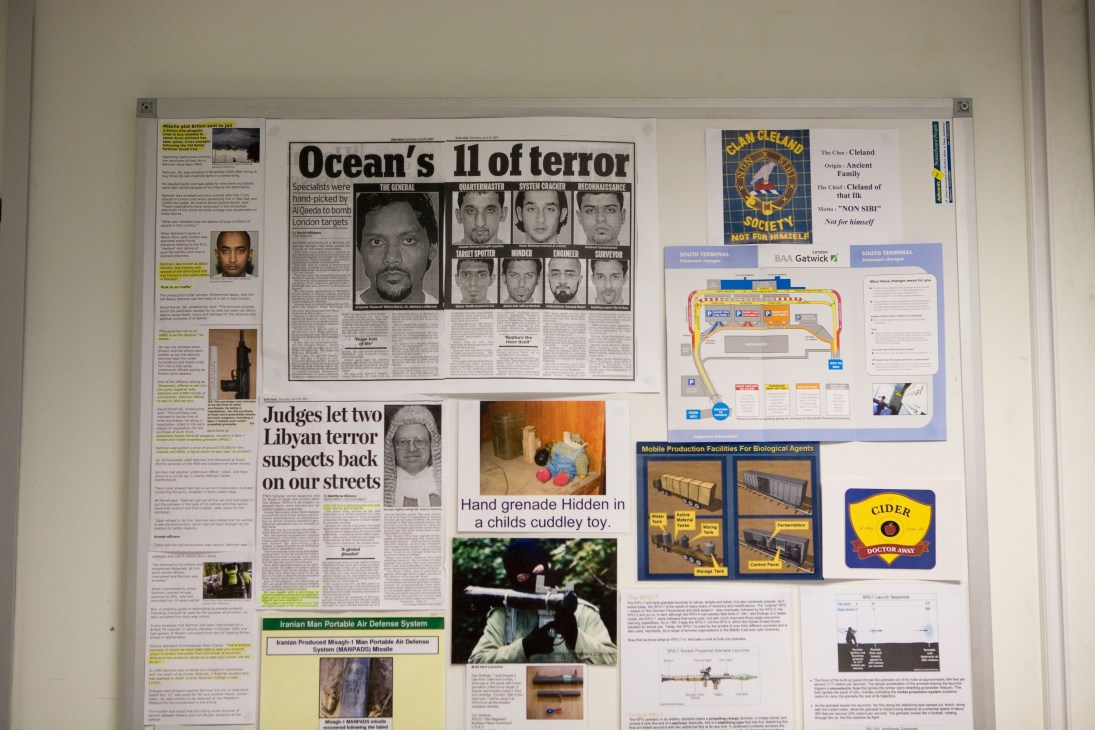 Security briefing board with newspaper clippings, threat intelligence photos, airport maps, and MANPAD system diagrams displayed on a wall.