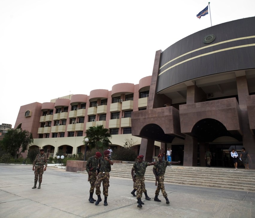Soldiers in camouflage uniforms stand in formation on a plaza outside a modern government building with pink and cream colored architecture and a Cape Verdean flag.