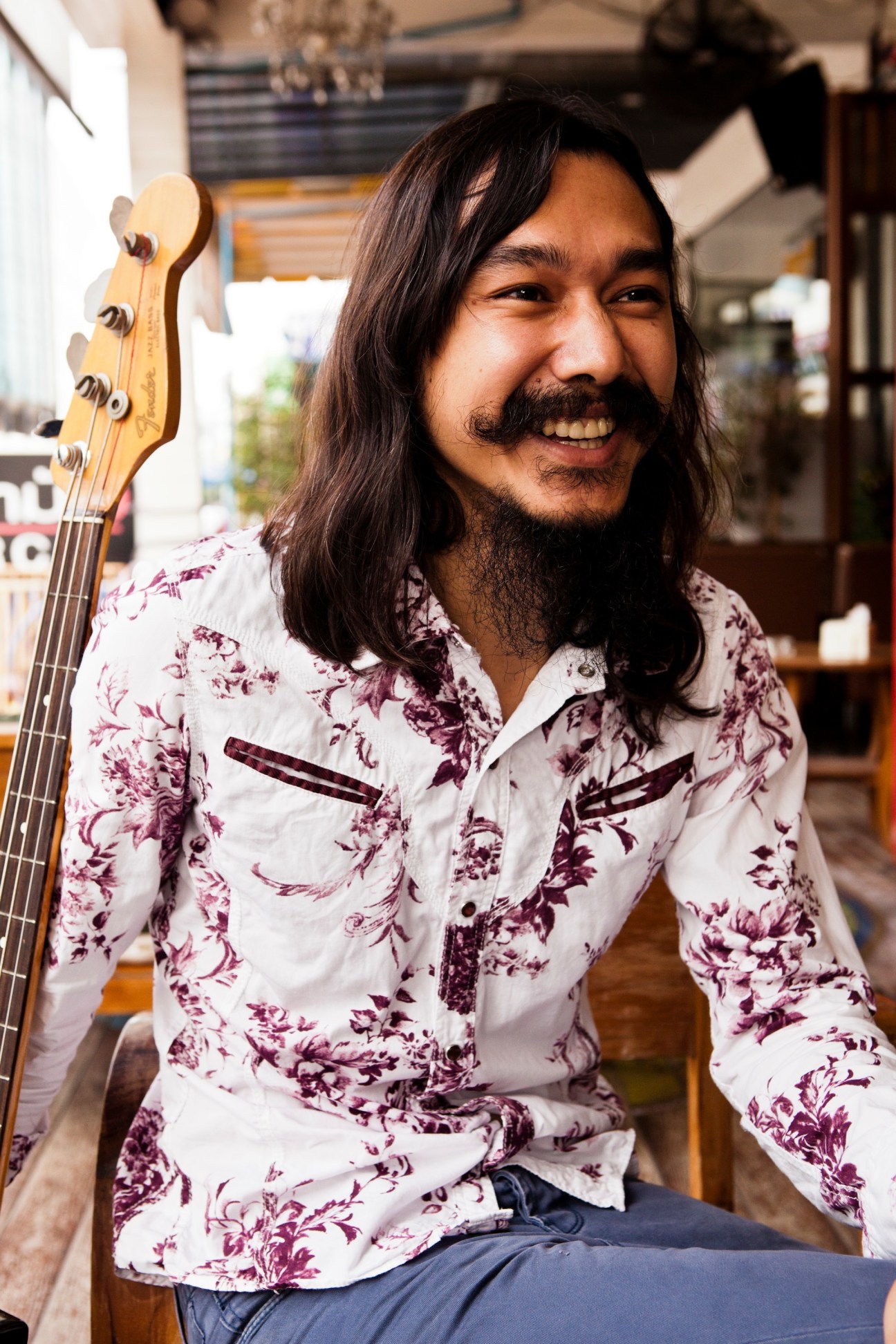 Man with long dark hair and mustache holding electric bass guitar, wearing floral patterned shirt at a café.