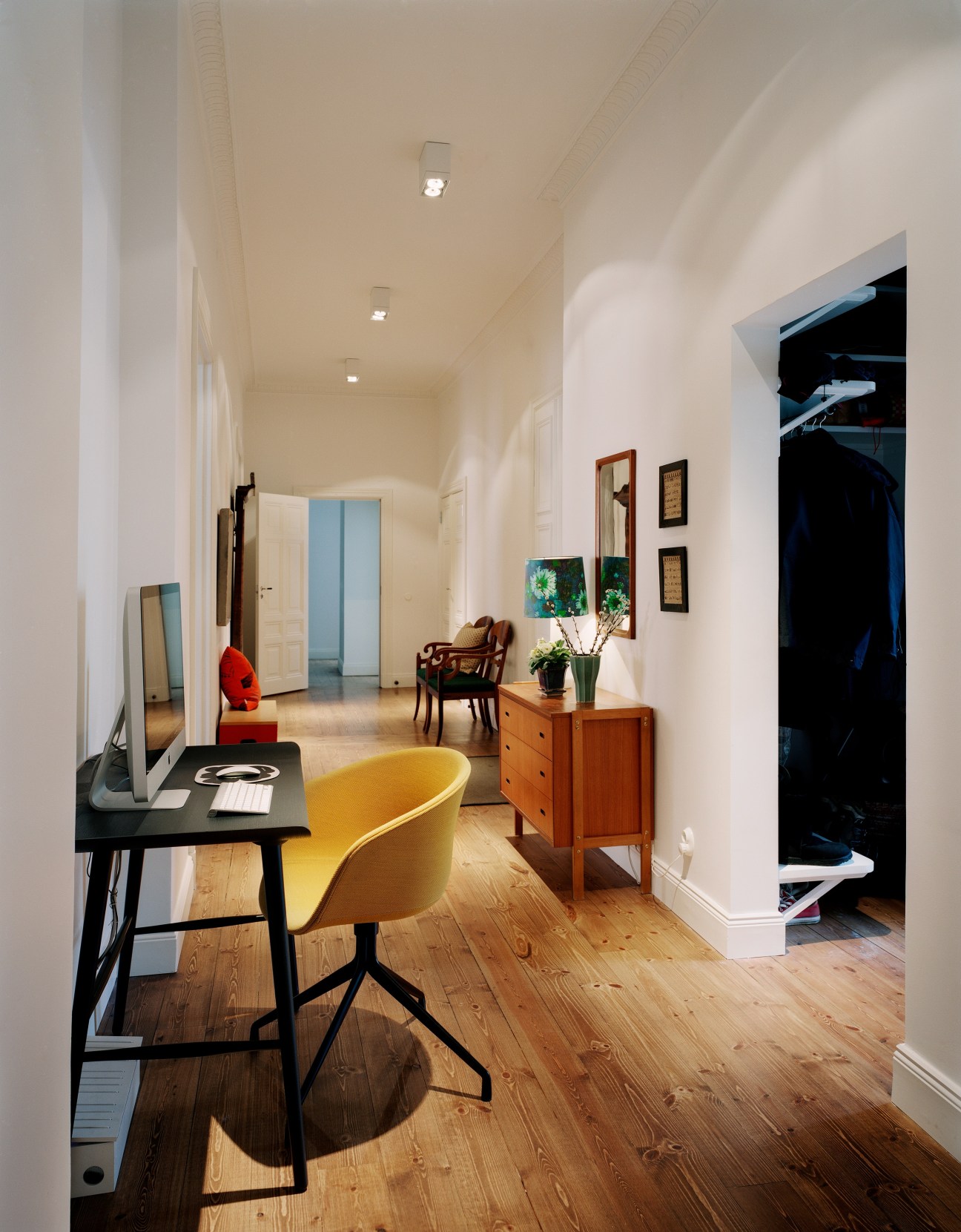 A bright hallway with white walls, wooden floors, and a yellow chair at a black desk used as a home office.