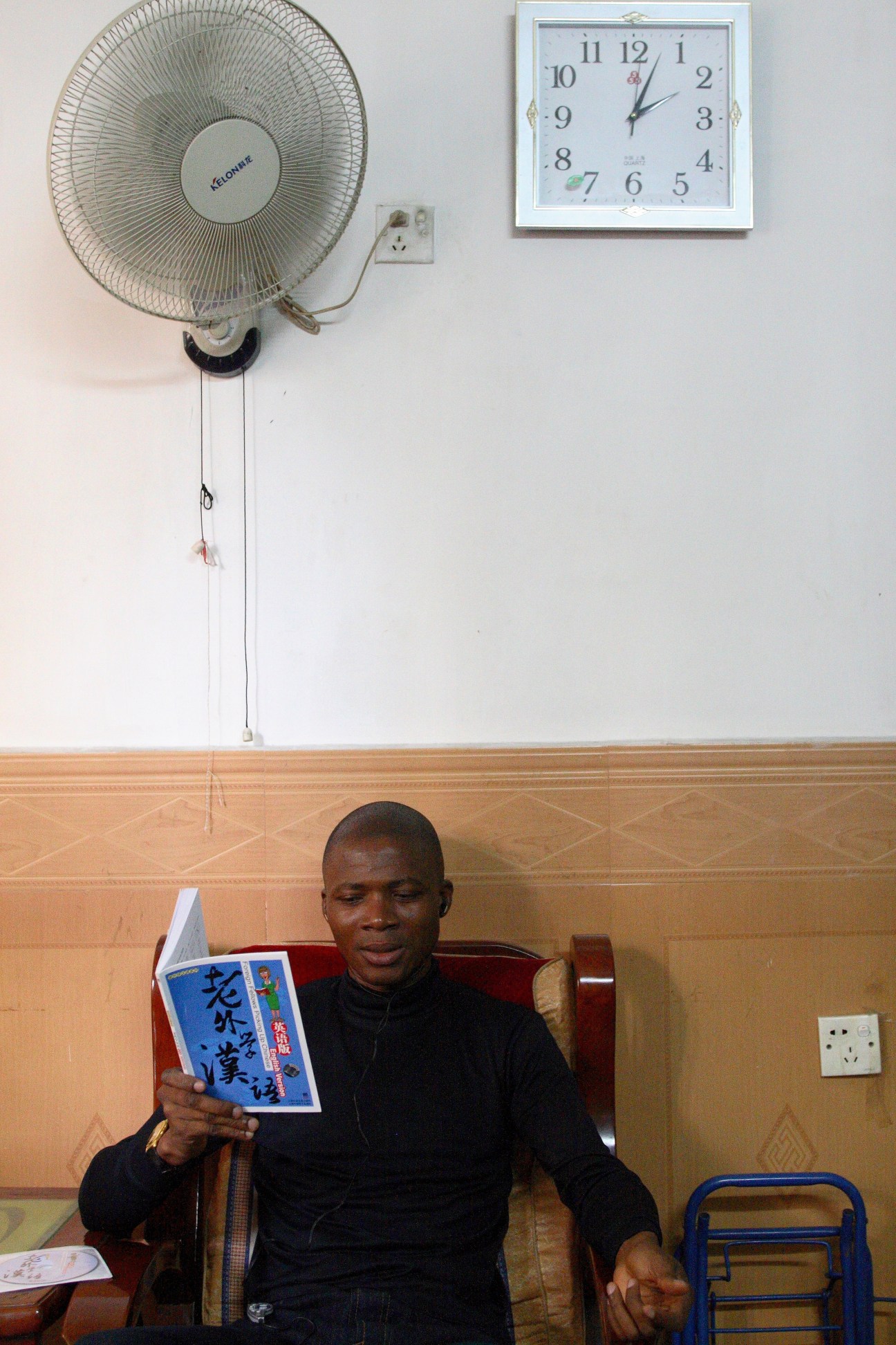Man seated indoors holding a Chinese language learning book beneath a wall-mounted fan and clock.