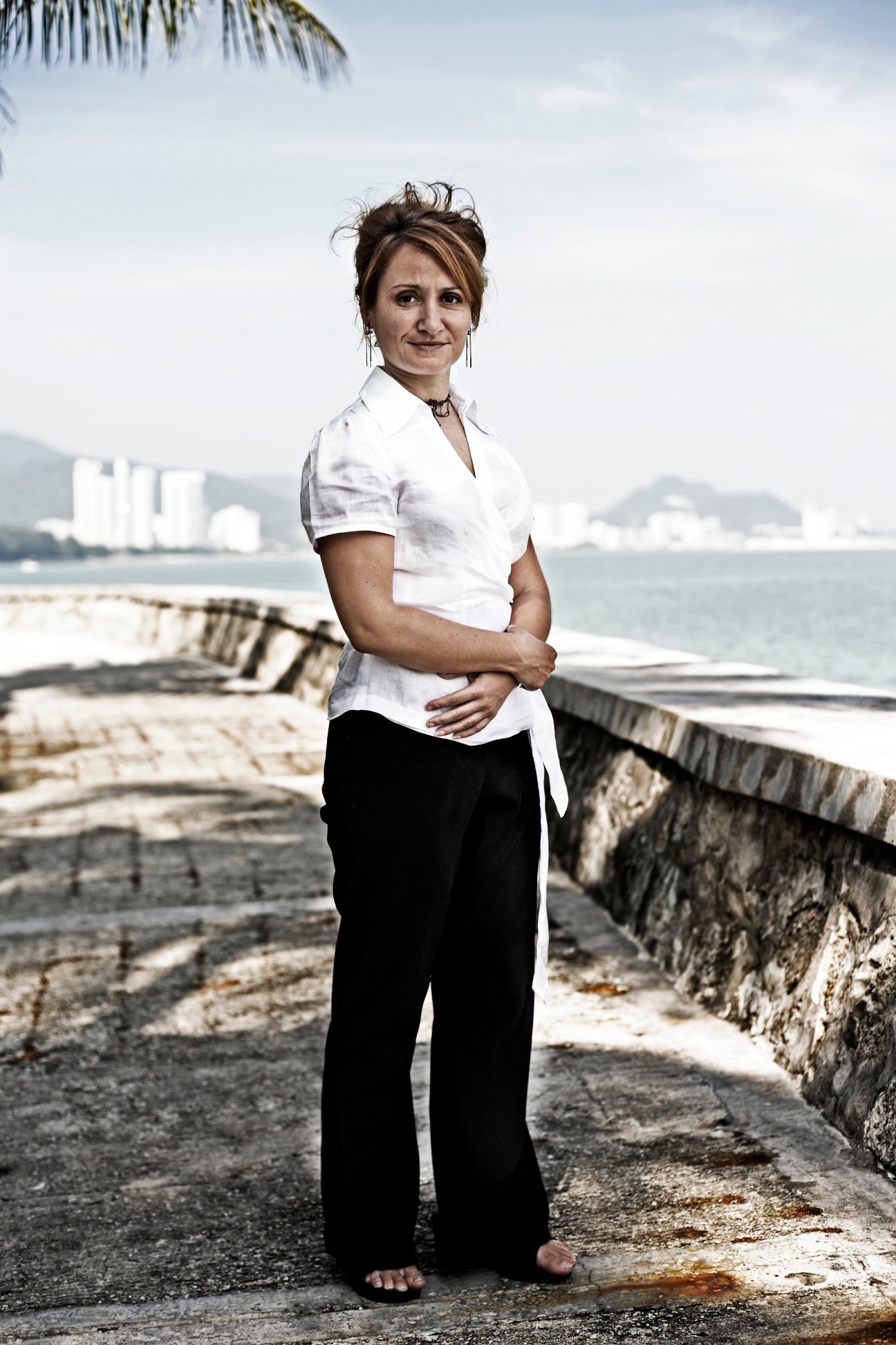 Michelle Peake stands on a waterfront promenade in George Town with colonial buildings and sea visible behind her.