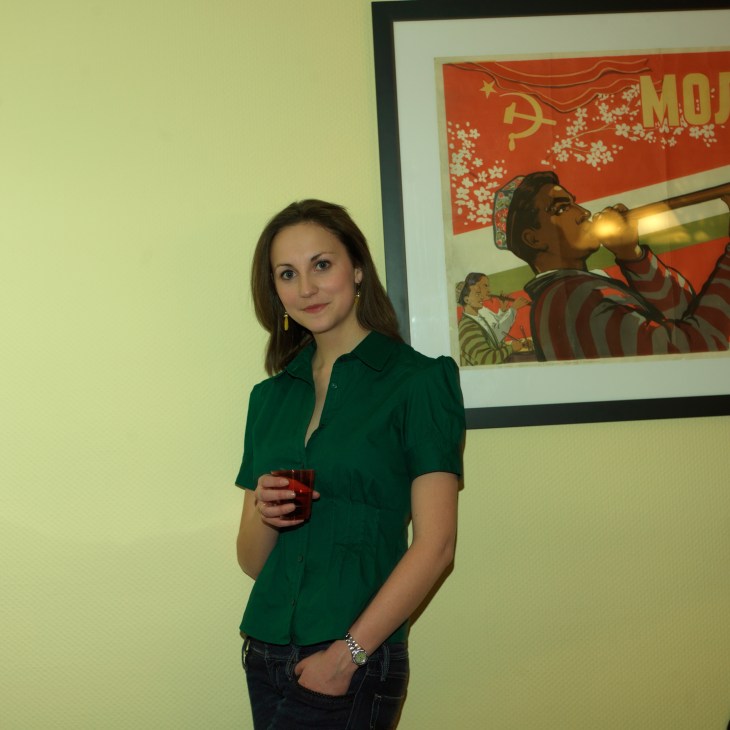 Woman in green shirt holding a red cup stands in front of Soviet propaganda poster.