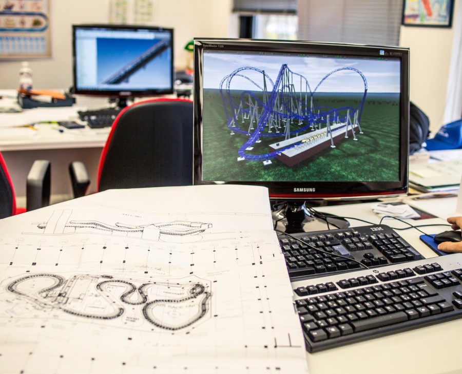 3D computer model of a blue roller coaster displayed on a Samsung monitor in a design office, with technical drawings visible on the desk.