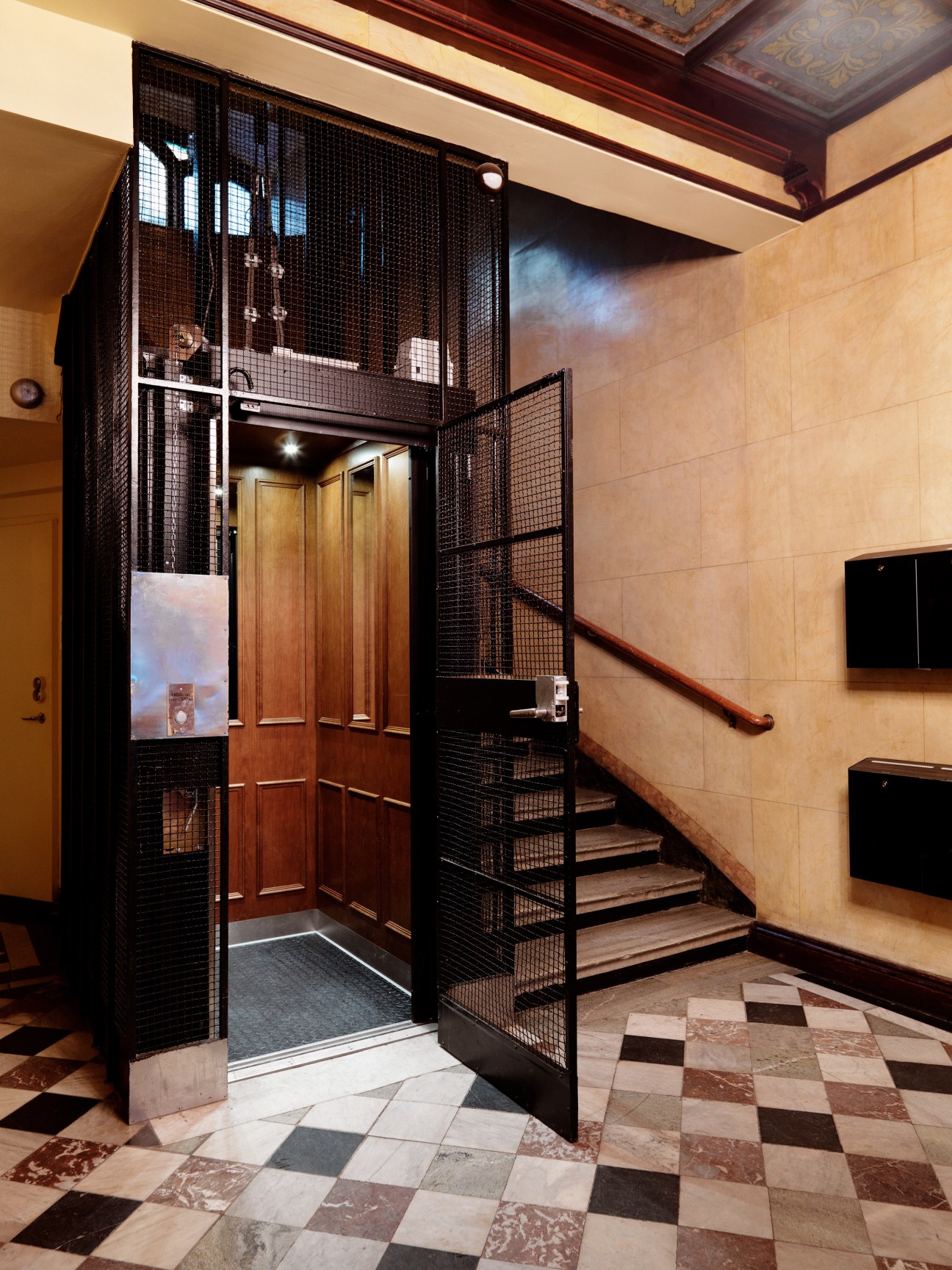 Wrought iron cage entrance to a wood-panelled lift in a stairwell with cream and terracotta tile flooring.