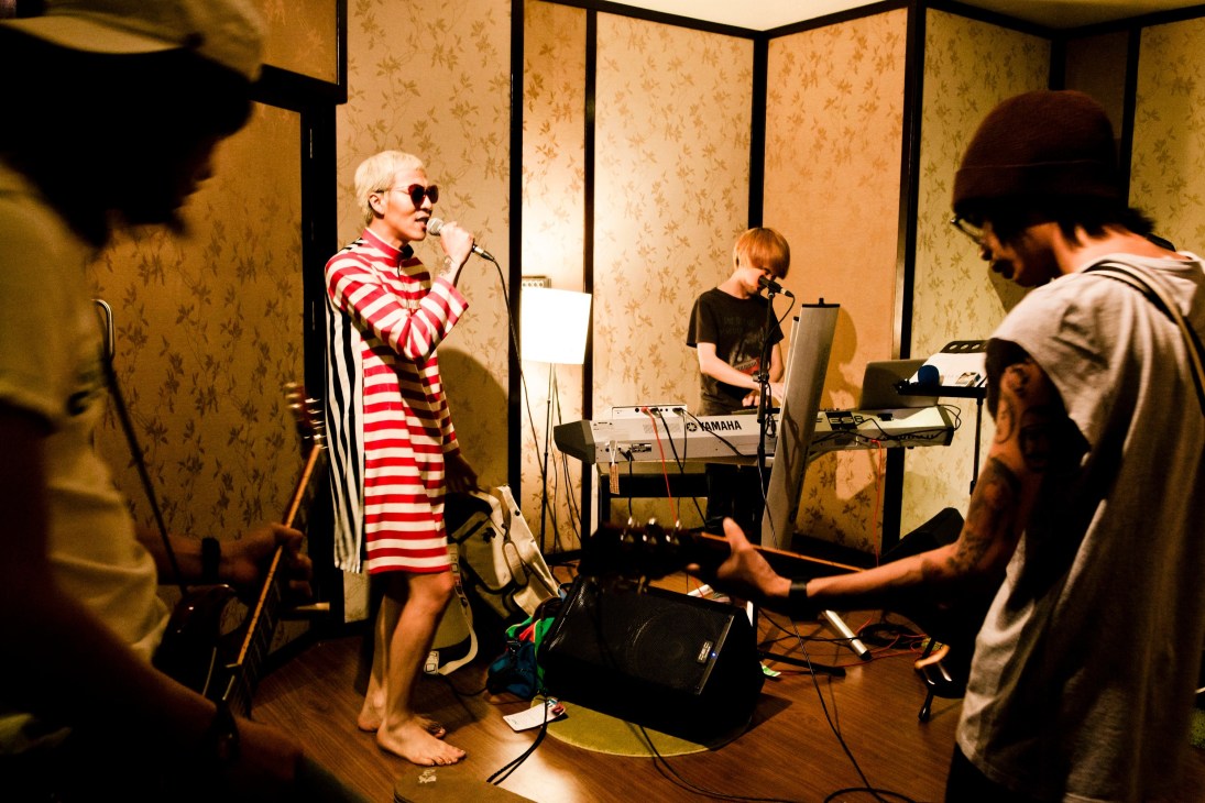 Gene Kasidit performs in red and white striped clothing with a band in a room with patterned wallpaper.