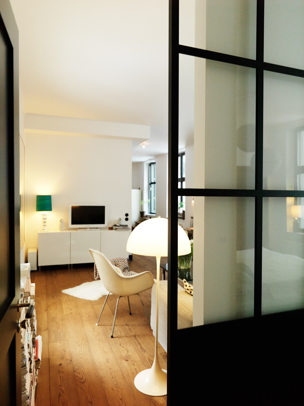 Modern bedroom separated from living area by black-framed glass sliding doors with wooden flooring.