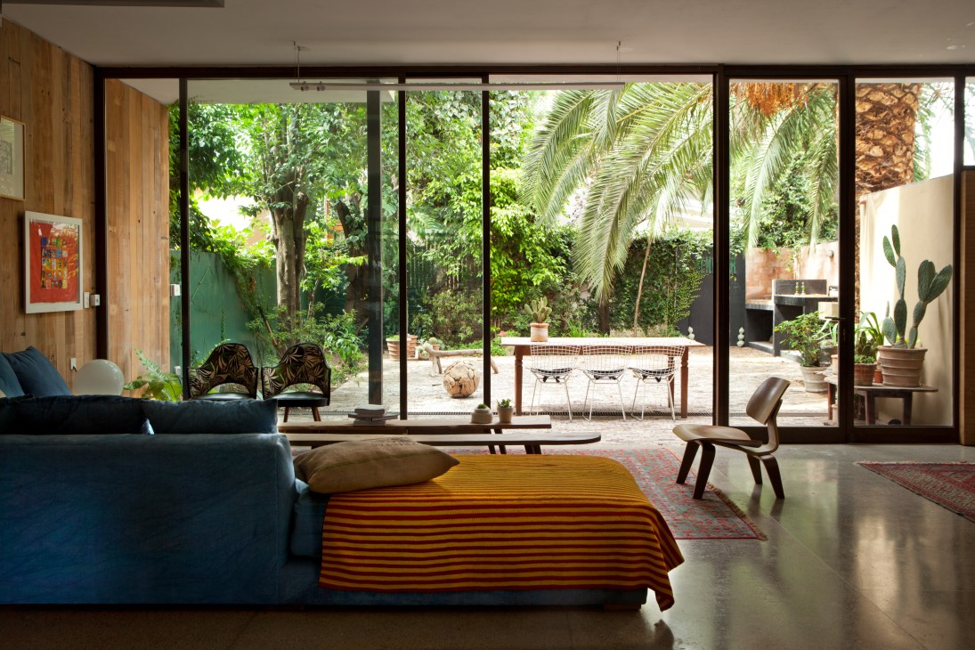 Open-plan living room with floor-to-ceiling windows overlooking a lush garden with palm tree and brick barbecue.