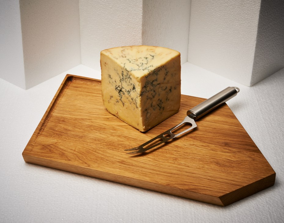 Oiled ash chopping board with blue cheese and metal serving fork on white surface.