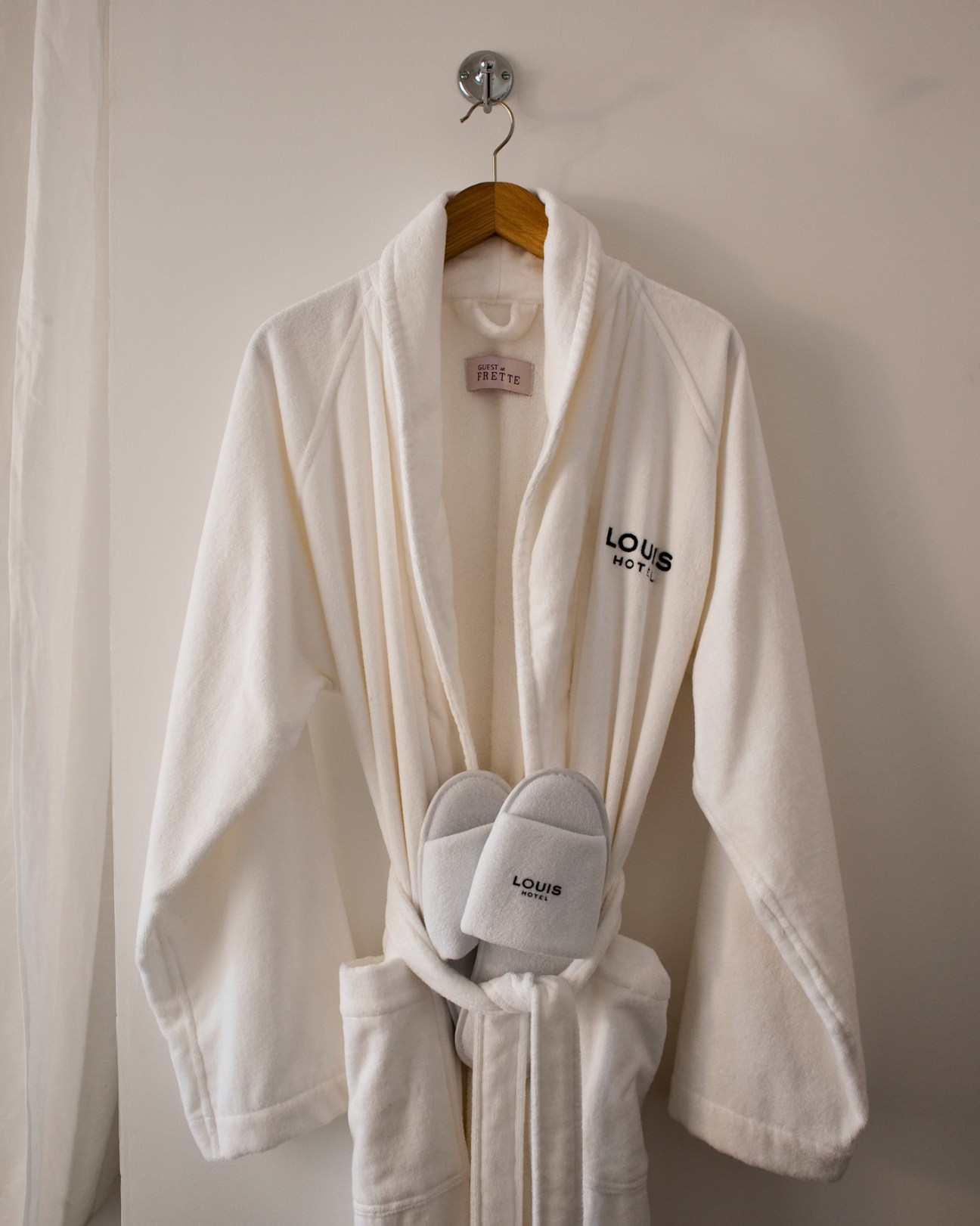 White Louis Hotel bathrobe and slippers hanging on wooden hanger against white wall.