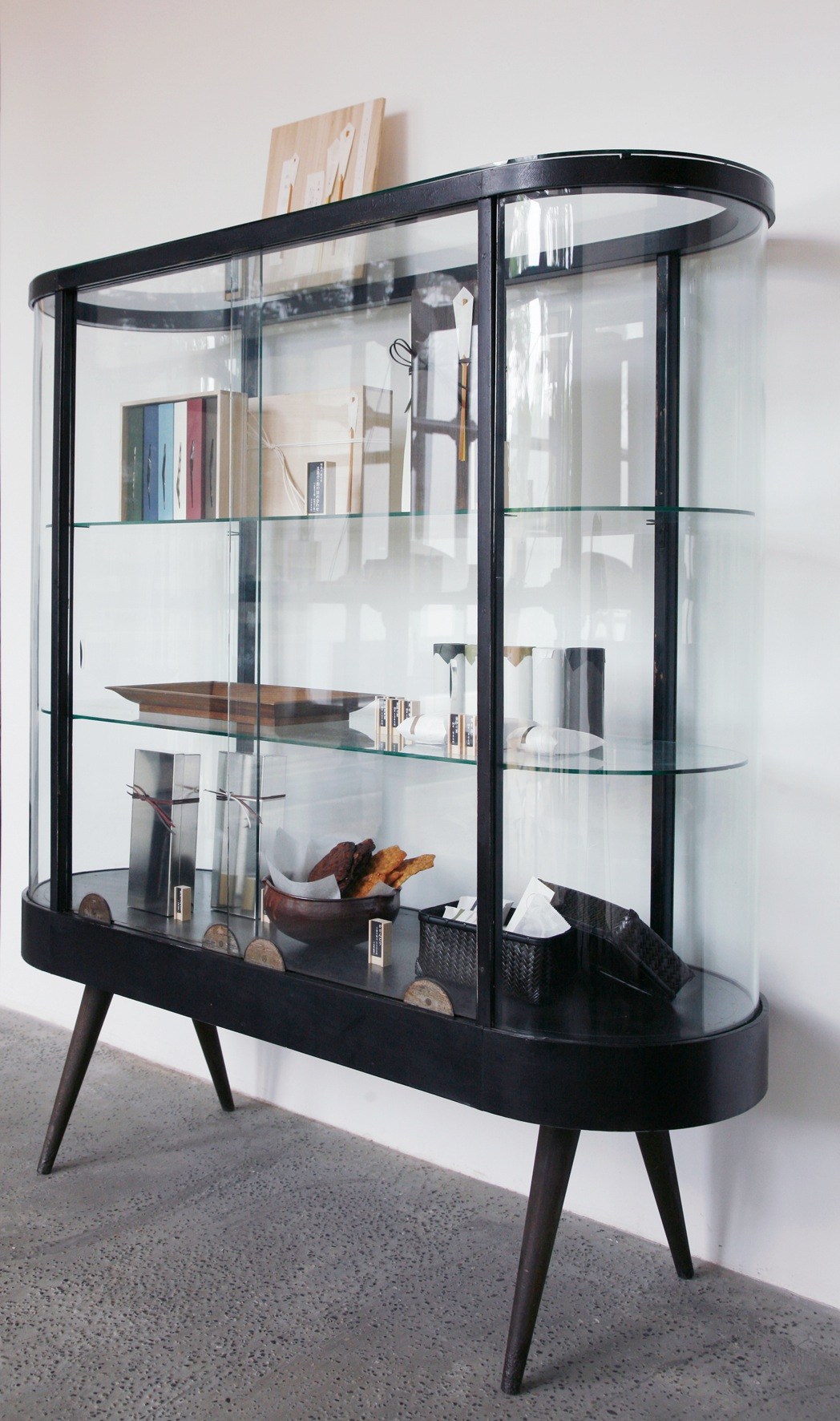 Mid-century modern display cabinet with curved black frame, glass panels, and tapered legs containing books, ceramics, and decorative objects.
