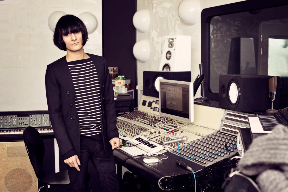 Thomas Troelsen stands in his studio surrounded by synthesisers and recording equipment, wearing a black blazer and striped shirt.