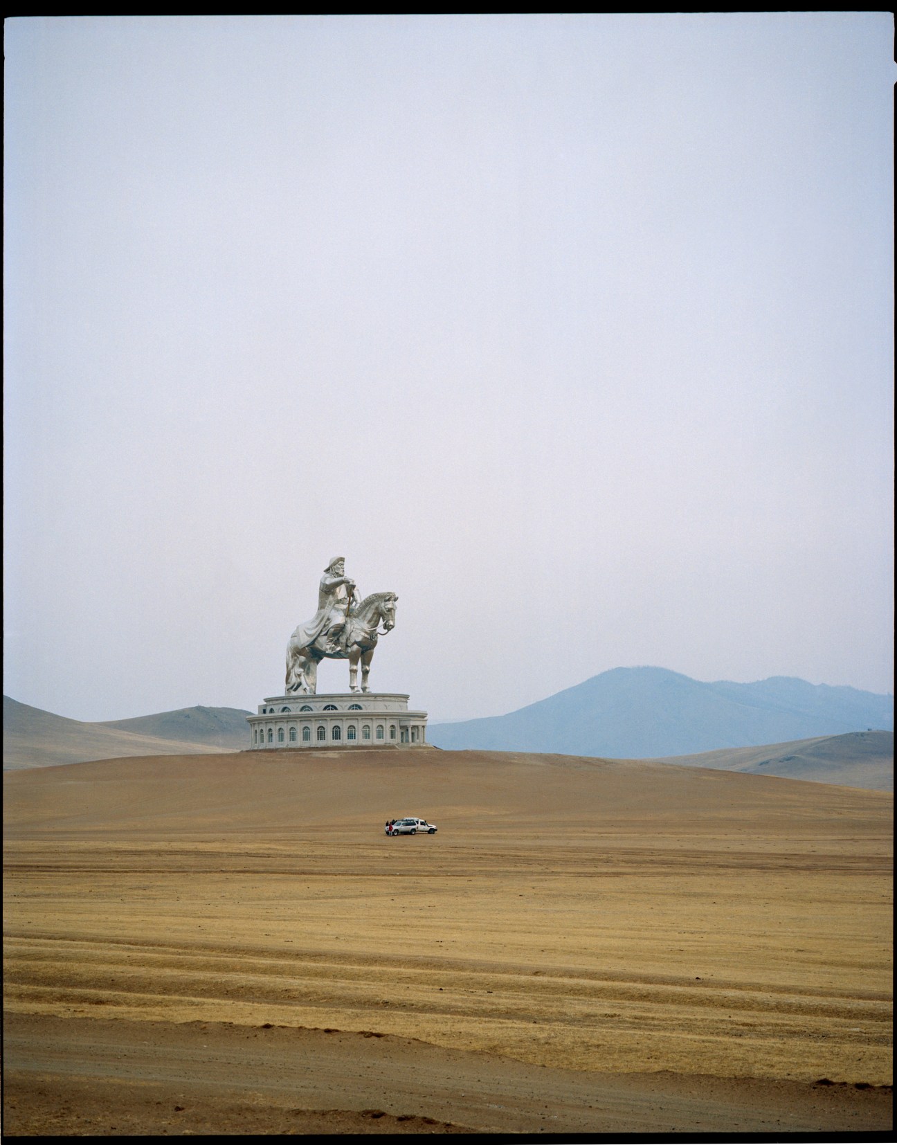 A 40-meter steel statue of Genghis Khan on horseback stands on a stone platform in a barren Mongolian landscape with mountains in the distance and a car driving across the steppe.