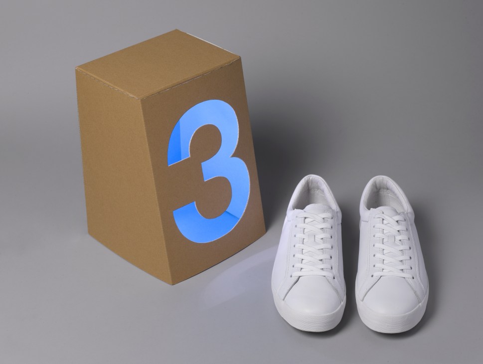 Pair of white canvas trainers displayed next to a cardboard box marked with a blue number 3.
