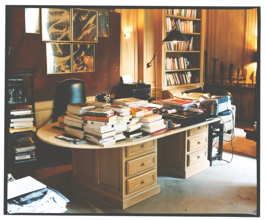 Wood-panelled office with a large desk piled with books, bookshelves, and warm lighting.