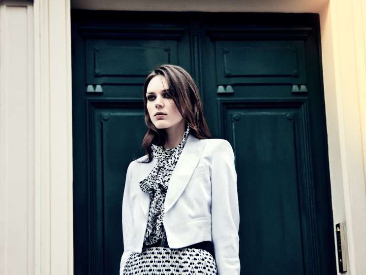 Model wearing white jacket by René Lezard and patterned dress by Oscar de la Renta stands before a green door numbered 12 in Paris.