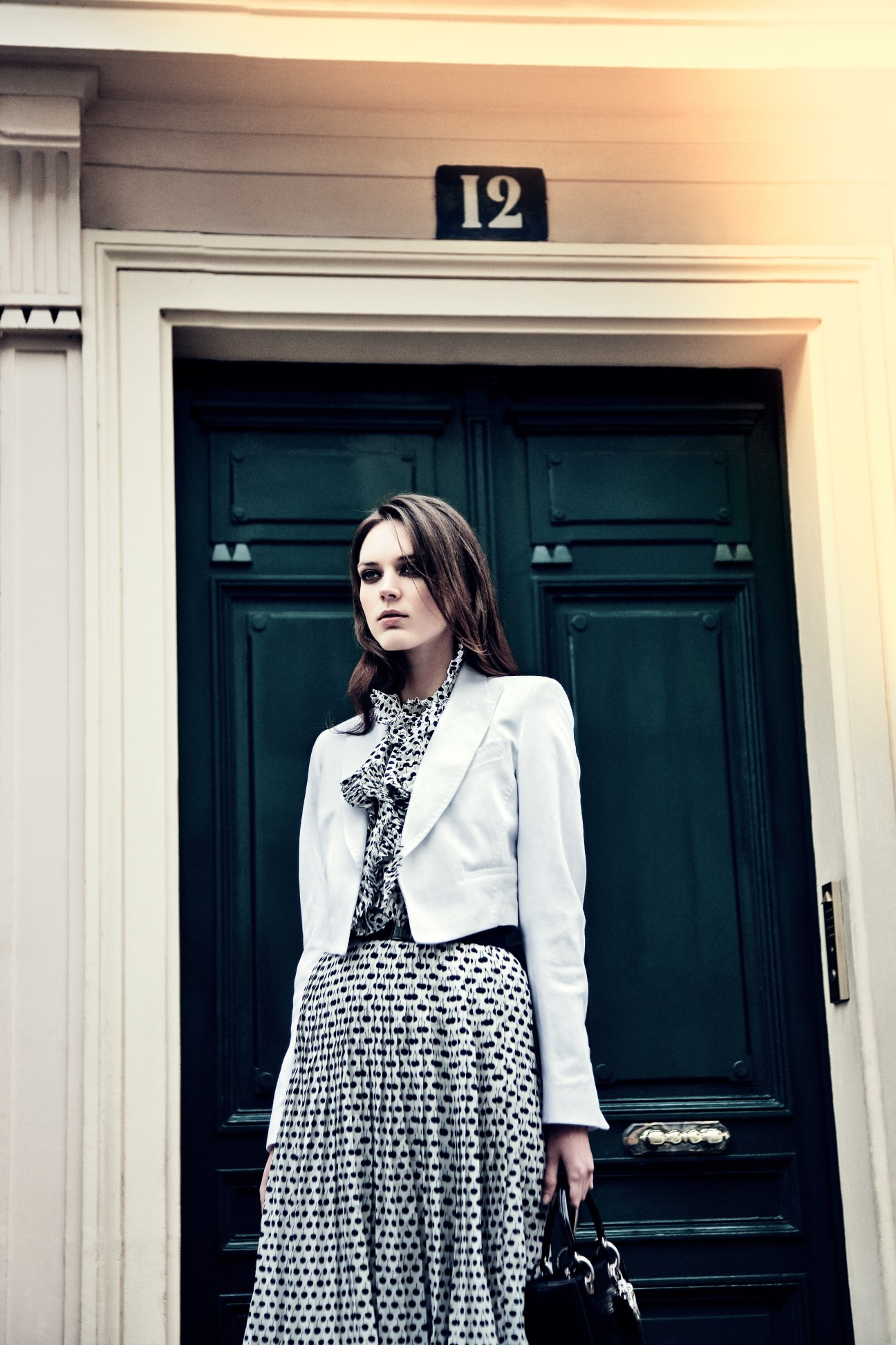 Model wearing white jacket by René Lezard and patterned dress by Oscar de la Renta stands before a green door numbered 12 in Paris.