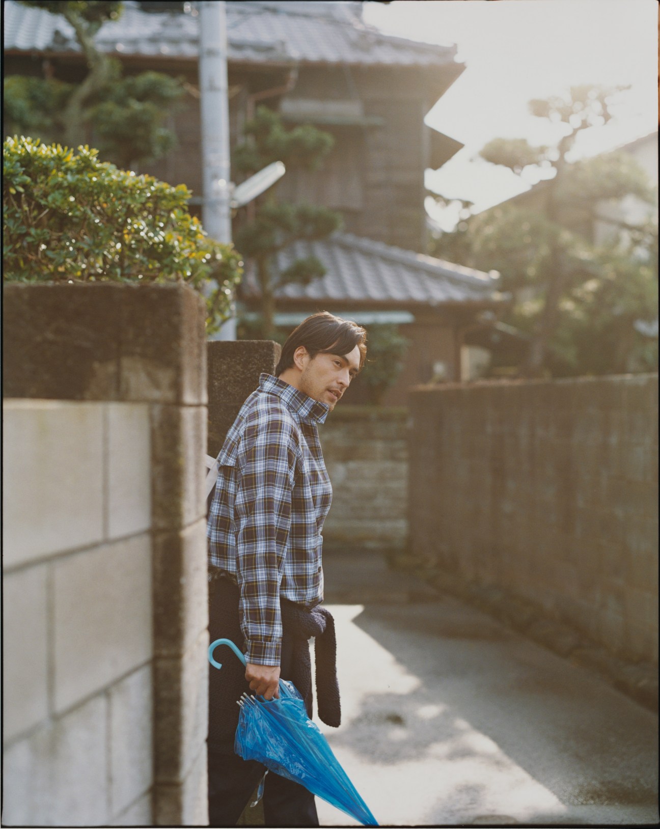 Man in blue plaid shirt and black trousers holding blue umbrella in Japanese residential alley.