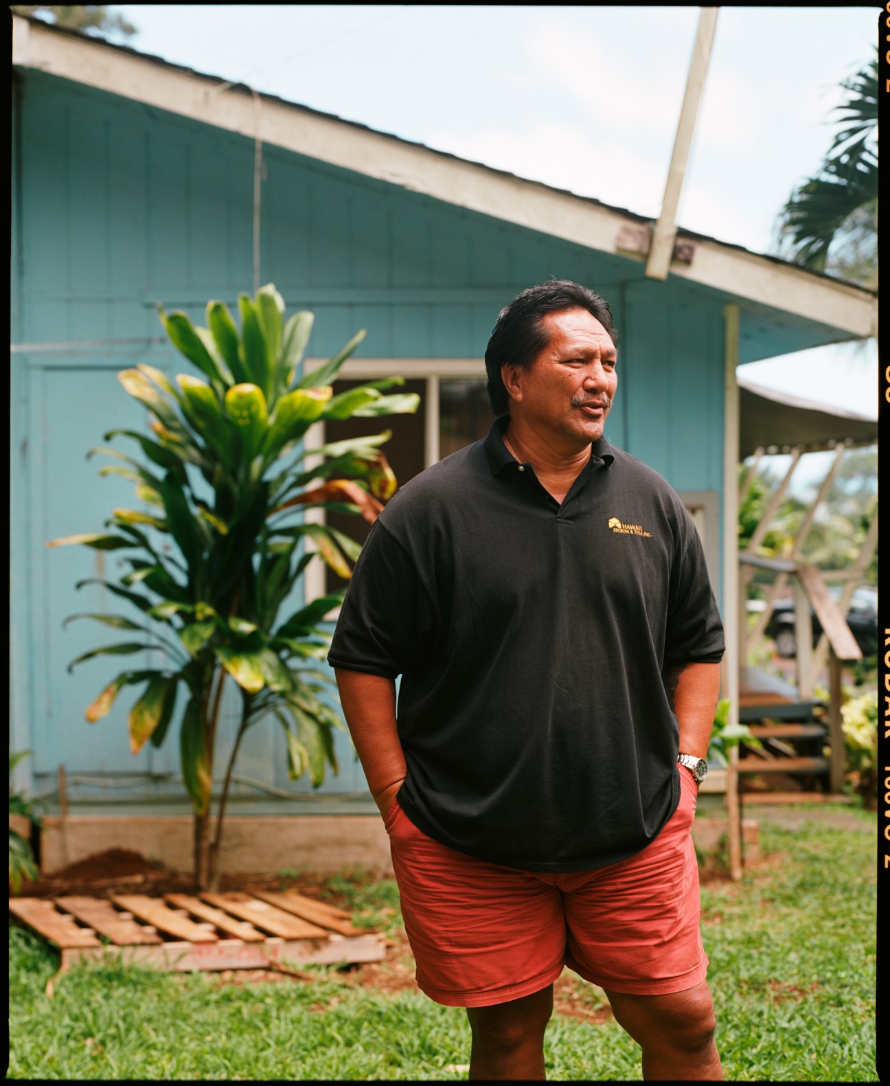 Bumpy Kanahele stands outside a simple building with a potted plant, wearing a black polo shirt and red shorts.